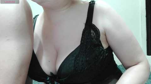 Video of a fair-skinned woman with large breasts wearing a black lace bra, partially visible. Background shows a white wall and a dark object.