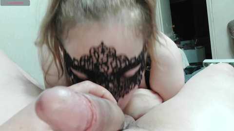 Video of a fair-skinned, blonde woman wearing a black lace mask, kneeling on a bed with her mouth close to a large erect penis. The background shows a dimly lit room with a doorway leading to another area.