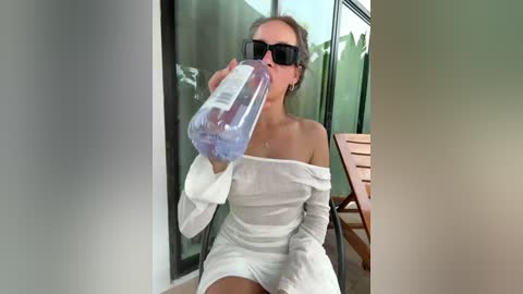 A video of a woman with light skin, wearing dark sunglasses, an off-shoulder white dress, and drinking from a plastic water bottle on a patio with glass doors, wooden chairs, and green plants in the background.
