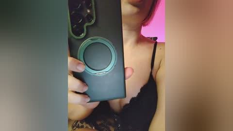 Video of a woman with fair skin, wearing a black lace bra, holding a green smartphone with a circular lens. Her shoulder and part of her face are visible. Background is pink and blurry.