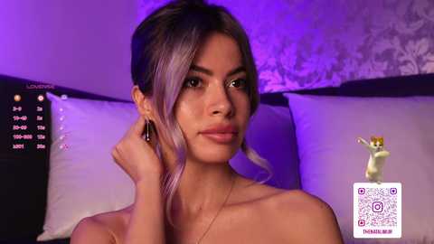 Video of a young woman with light skin and long, dark hair styled in a side part, wearing a gold necklace. She sits on a bed with white pillows and a dark headboard, bathed in purple lighting.