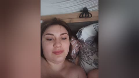 A video of a light-skinned woman with medium-length brown hair, wearing a neutral expression, lying on a bed with patterned pillows. Background shows a wooden headboard with a black spider figurine.