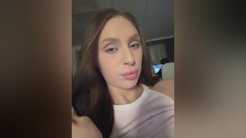 Video of a young woman with fair skin, straight brown hair, and light makeup, wearing a white shirt. She's in a dimly lit room, possibly a bedroom, with blurred background elements.