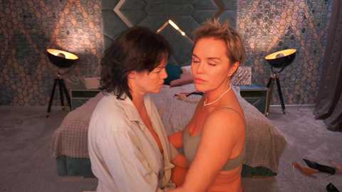 Video of two women, one in a white shirt, the other in a green bra, in a dimly lit, ornate bedroom with patterned walls and a bed.
