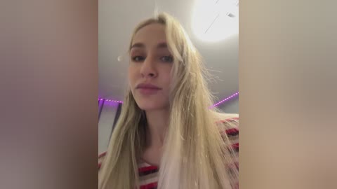 Video of a young, fair-skinned woman with long, straight blonde hair, wearing a red and white striped top. She has a neutral expression and is indoors with a purple LED strip lighting in the background.