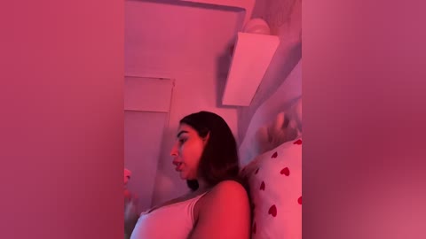 Video of a woman with dark hair, wearing a white top, leaning against a white bed with red heart pillows, bathed in pink lighting, creating a sensual, intimate atmosphere.