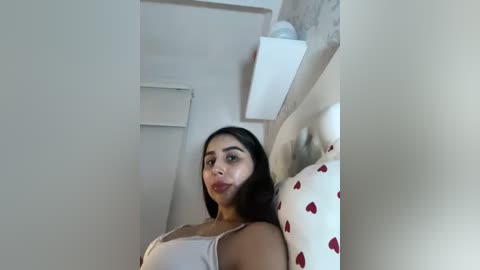 Video of a young woman with long black hair, medium skin tone, wearing a light gray tank top, standing in a small, white room with a white headboard and heart-patterned pillow.