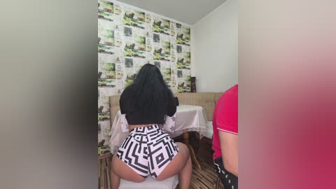 Video of a woman in black clothing, seated on a white table, facing a newspaper-patterned wall, with a red-shirted person partially visible in the background.