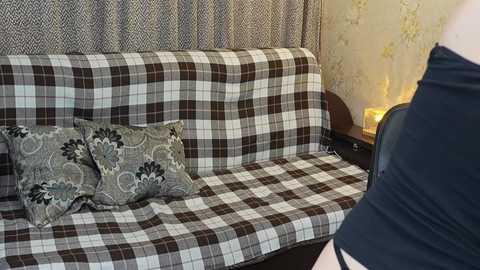 Video of a cozy living room with a plaid-patterned couch, floral pillows, and a person wearing black clothing, against a wallpapered backdrop with floral designs.