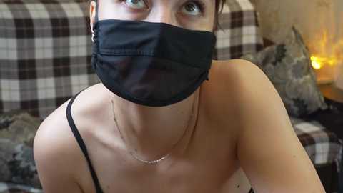 Video of a light-skinned woman with short brown hair, wearing a black face mask and black spaghetti strap top, sitting on a checkered couch in a dimly lit room.