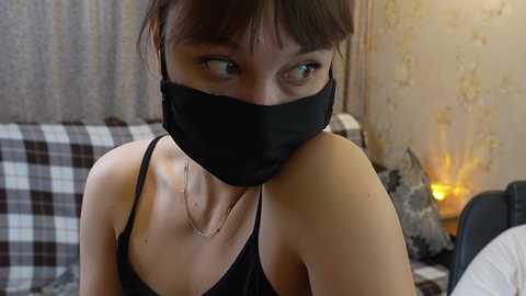 A video of an Asian woman with light skin, wearing a black face mask, black tank top, and a delicate necklace, sitting indoors with a plaid-patterned bedspread and a lit candle in the background.