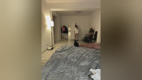 A video of a dimly-lit, sparse bedroom with a man lying on a gray blanket-covered bed, a woman in a white dress standing in the corner, and a large, soft, beige lampshade casting a warm glow.