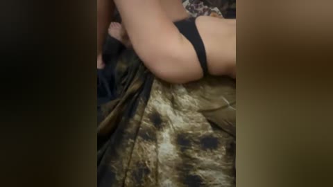 Video of a partially visible, light-skinned person with a slender build, wearing a black bra, lying on a bed with a patterned green and brown comforter. The image is blurred and grainy.