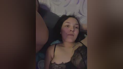 A video captures a young Asian woman with medium skin tone and black hair, lying on a bed with a floral-patterned pillowcase. She wears a black lace bra, and her expression is neutral. The image is slightly blurry.