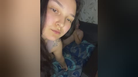 A video of a young woman with medium skin tone, long dark hair, and a tattoo on her upper arm, lying on a bed with a blue-patterned blanket.