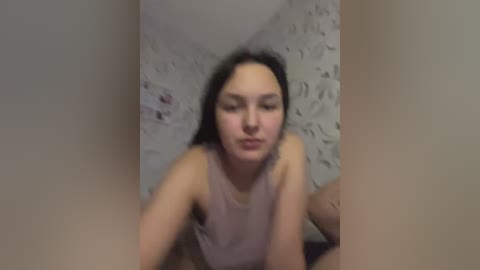 A blurry video of a young woman with light skin, long black hair, and a pink tank top, sitting on a bed with floral-patterned sheets.