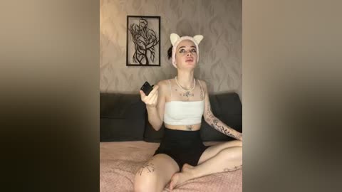 Video of a pale-skinned woman with black hair, wearing a white bandeau top, black shorts, and white cat ears, sitting on a bed with a pink blanket.