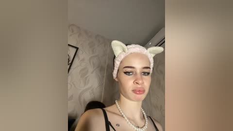 Video of a fair-skinned woman with blue eyes, wearing a white headband with bunny ears, a pearl necklace, and black top. She stands indoors, with a patterned wallpaper and a framed picture in the background.