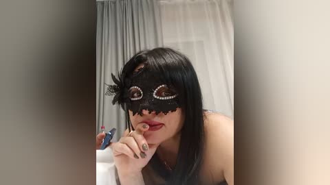 Video of a young woman with straight black hair, wearing a black lace masquerade mask with intricate detailing and a feather, holding a blue marker near a white curtain.