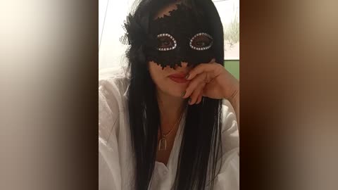 Video of a woman with long black hair, wearing a black lace masquerade mask with large, circular eye openings and a floral design. She has a light skin tone, red lipstick, and is dressed in a white robe. The background is blurred.