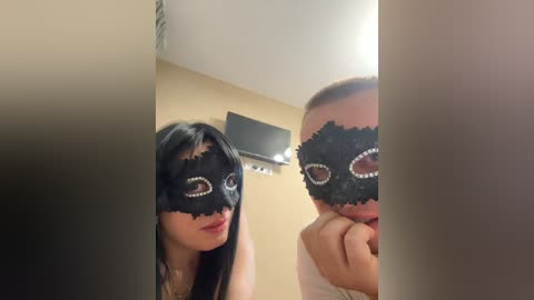 Video of two people in a room wearing black, lace Venetian masks with red eye holes. The woman on the left has long black hair, while the man on the right has short hair and a light skin tone.