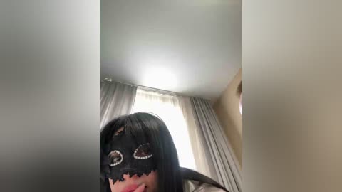 A video shows a person with long black hair, wearing a black mask with large, white, cat-like eyes, standing in a dimly lit room with white curtains and beige walls.
