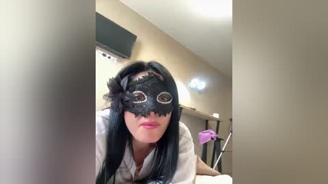 Video of a young woman with long black hair, wearing a black lace masquerade mask with glittery eyes, and a white shirt. She is indoors, in a room with beige walls and a TV mounted on the wall.