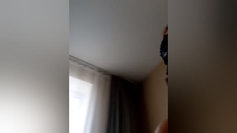 Video of a person with light skin and short hair, partially hidden behind a beige wall, wearing dark clothing, in a dimly lit room with white curtains and a window.