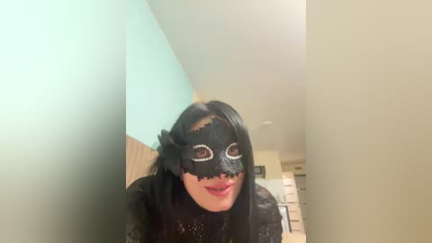 Video of a woman with long black hair, wearing a black lace mask with white polka dots and a black feathered headband, smiling against a light blue wall in a modern, minimalist room.