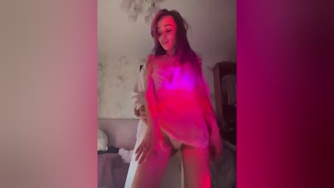 Video of a woman with long brown hair in a white shirt, standing in a dimly lit room with pink and purple light, revealing her breasts and genitals.