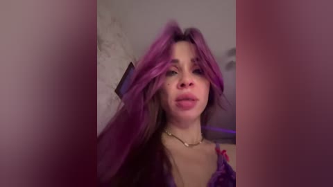 Video of a young woman with purple hair, wearing a pink lace bra, standing in front of a textured wall, with a blurred background.