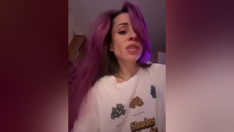 A video of a woman with vibrant purple hair, wearing a white shirt with colorful animal logos, captured indoors with a purple and red blurred background.