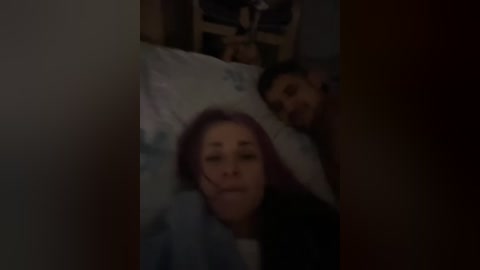 A dimly lit video shows a woman with light skin and shoulder-length red hair lying on a bed, partially obscured by a pillow, with a man's head visible behind her. The room has a wooden nightstand with a lamp and a white pillowcase.