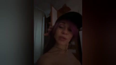 A dimly lit video of a topless woman with long dark hair and light skin, standing in a narrow doorway, partially obscured by shadows.