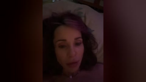 A video of a woman with curly hair, lying on a bed with dim lighting, partially covered by a blanket, her eyes closed, in a relaxed state.