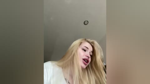 Video of a blonde woman with long hair, wearing a white top, mouth open, against a plain beige wall with a small hole.