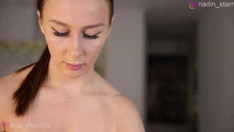 Video of a fair-skinned woman with long brown hair tied back, wearing dramatic black eyeliner and mascara, looking downward, with a blurred, neutral-toned background.