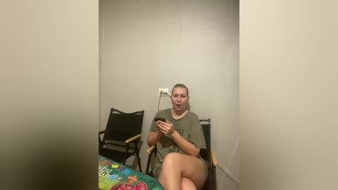 Video of a woman with short blonde hair, wearing a green T-shirt and shorts, sitting on a black folding chair in a plain room. She holds a white remote control, looking at it.