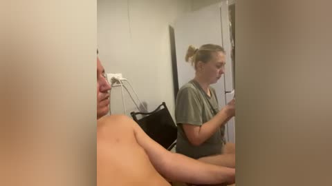 Video of a light-skinned woman in a green t-shirt, standing in a hospital room with a light-skinned, shirtless man in bed. The room has beige walls, a black bag, and a white electrical outlet.