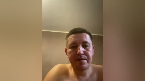 Video of a topless man with short dark hair and a round face, standing against a beige wall with a slight shadow. The man's expression is neutral.