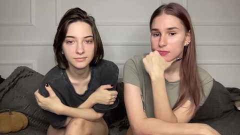 Video of two young women with fair skin, sitting on a bed with gray pillows, wearing casual outfits. One has dark hair, the other has long brown hair, both with thoughtful expressions.