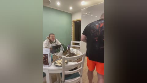 A video shows a white woman with light skin, short hair, wearing a beige sweater, seated at a white table with a brown bag and books, in a modern, green-painted room with wooden floors and recessed lighting.