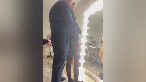 A video of a woman with long blonde hair, dressed in a blue dress, standing in a dressing room with a large mirror and bright lights.