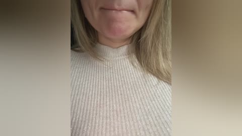 Video of a light-skinned woman with straight, shoulder-length blonde hair, wearing a ribbed, cream-colored turtleneck sweater. Her face is partially visible, showing a subtle, closed-mouth smile. The background is a plain, beige wall.
