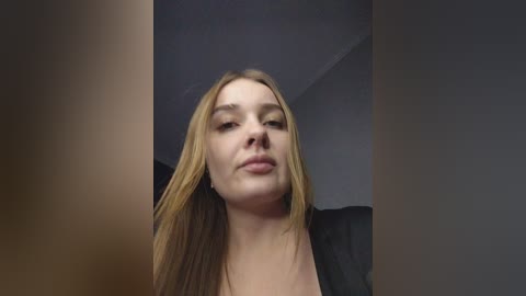 A close-up video of a young Caucasian woman with long, straight blonde hair, light skin, and a neutral expression, wearing a black top. The background is indistinct, possibly indoors with muted lighting.