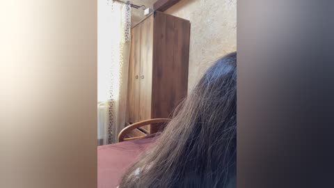 A video showing a person with long, dark hair lying on a bed in a dimly lit room. The background includes a wooden wardrobe and a window with sheer curtains.