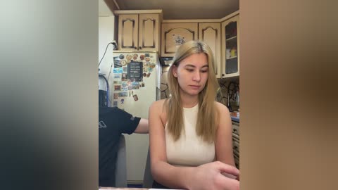 Video of a blonde Caucasian woman in a beige sleeveless top, sitting in a cluttered kitchen with wooden cabinets, refrigerator covered in magnets, and a black shirt hanging on a door.