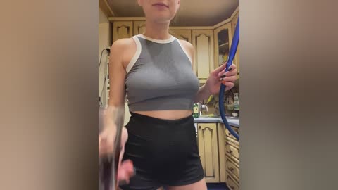 Video of a young woman in a tight, grey crop top and high-waisted black shorts, standing in a kitchen with wooden cabinets and a blue rope in her hand.