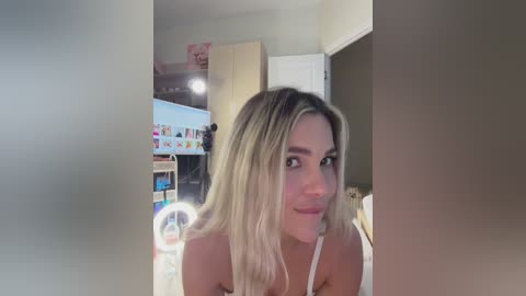 Video of a young woman with long blonde hair, fair skin, and a small tattoo on her shoulder, smiling slightly. Background includes a laundry room with a white washer and dryer, colorful artwork, and a closed door.