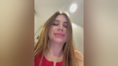 A video of a young woman with long, straight, light brown hair and fair skin, wearing a red top, smiling with eyes closed, standing indoors with a blurred background.